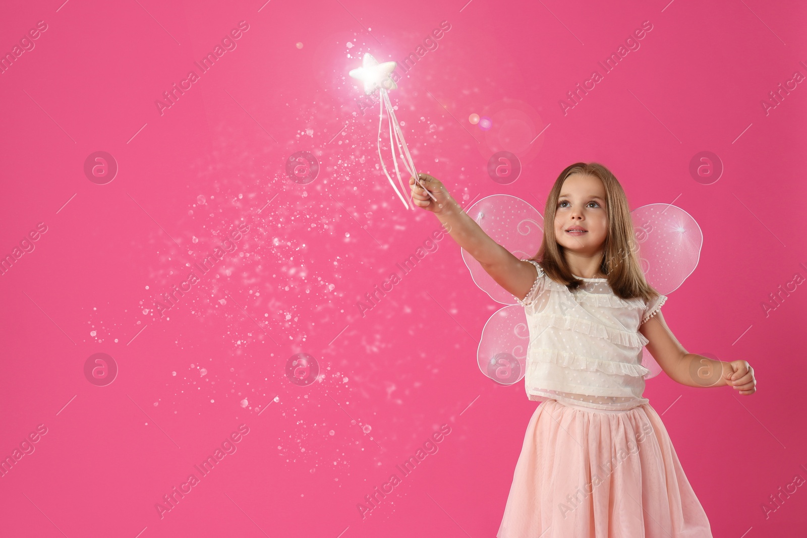 Image of Cute girl with fairy wings conducting spell on pink background. Magic lights around