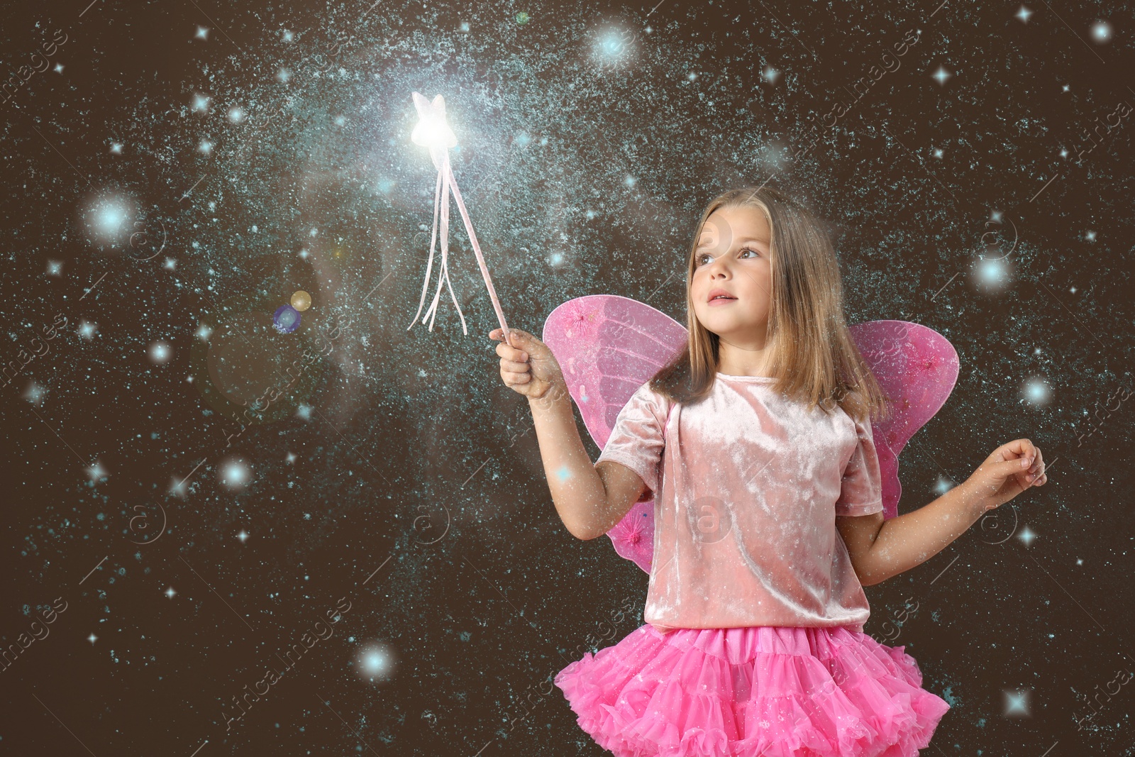 Image of Cute girl with fairy wings conducting spell on brown background. Magic lights around