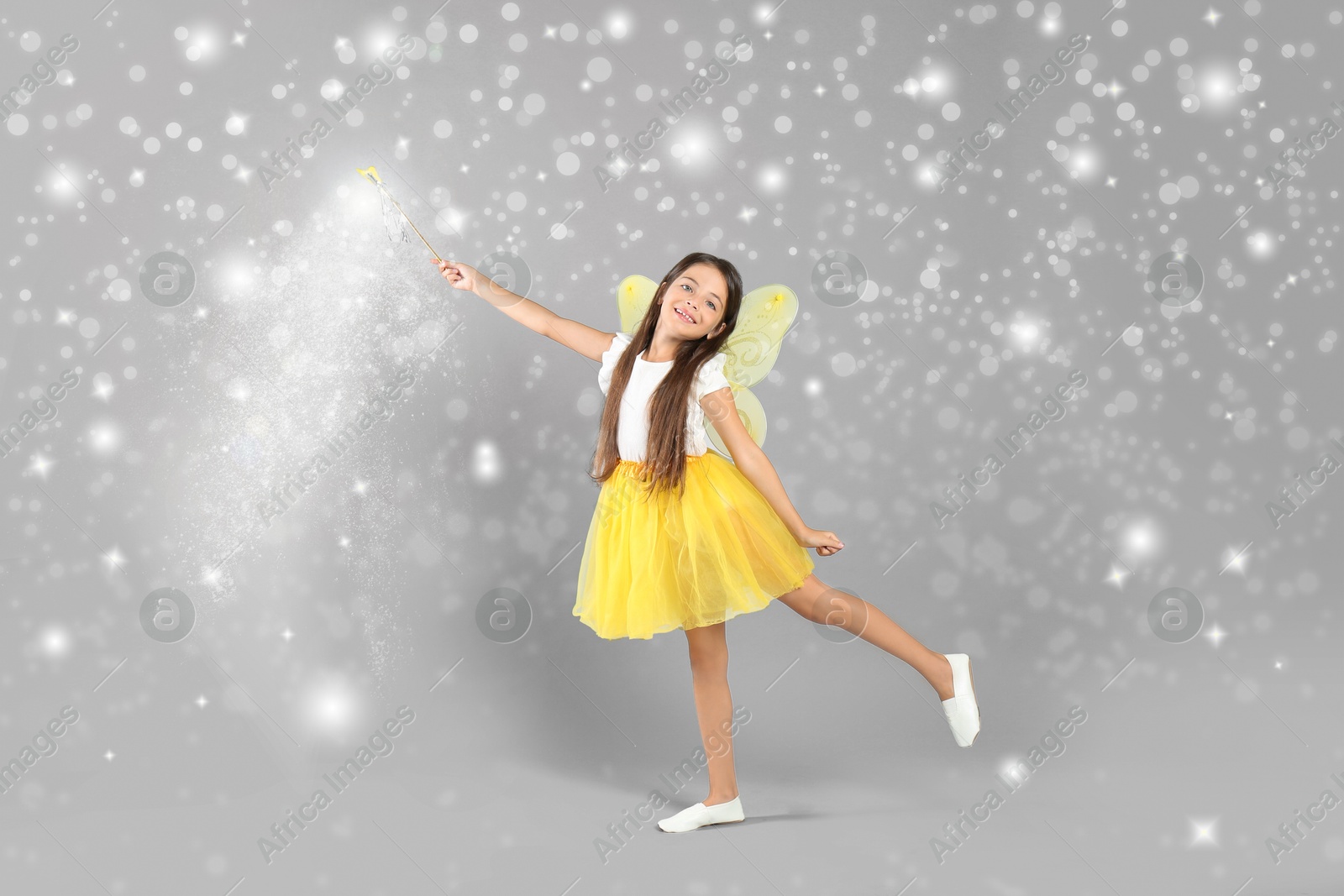 Image of Cute girl with fairy wings conducting spell on grey background. Magic lights around