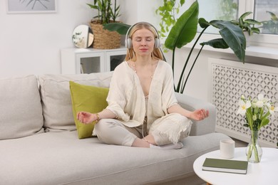 Photo of Feng shui. Young woman with headphones meditating on couch at home