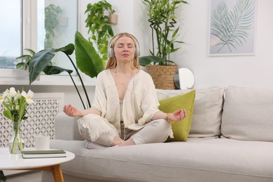 Photo of Feng shui. Young woman with headphones meditating on couch at home