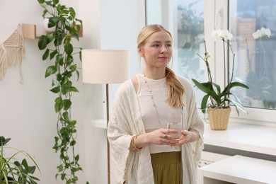 Photo of Feng shui. Young woman with glass of water near houseplants at home