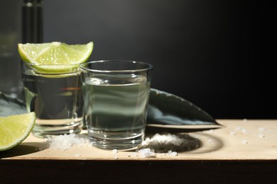 Photo of Tequila shots, slices of lime, salt and agave leaves on wooden table against black background, closeup. Space for text