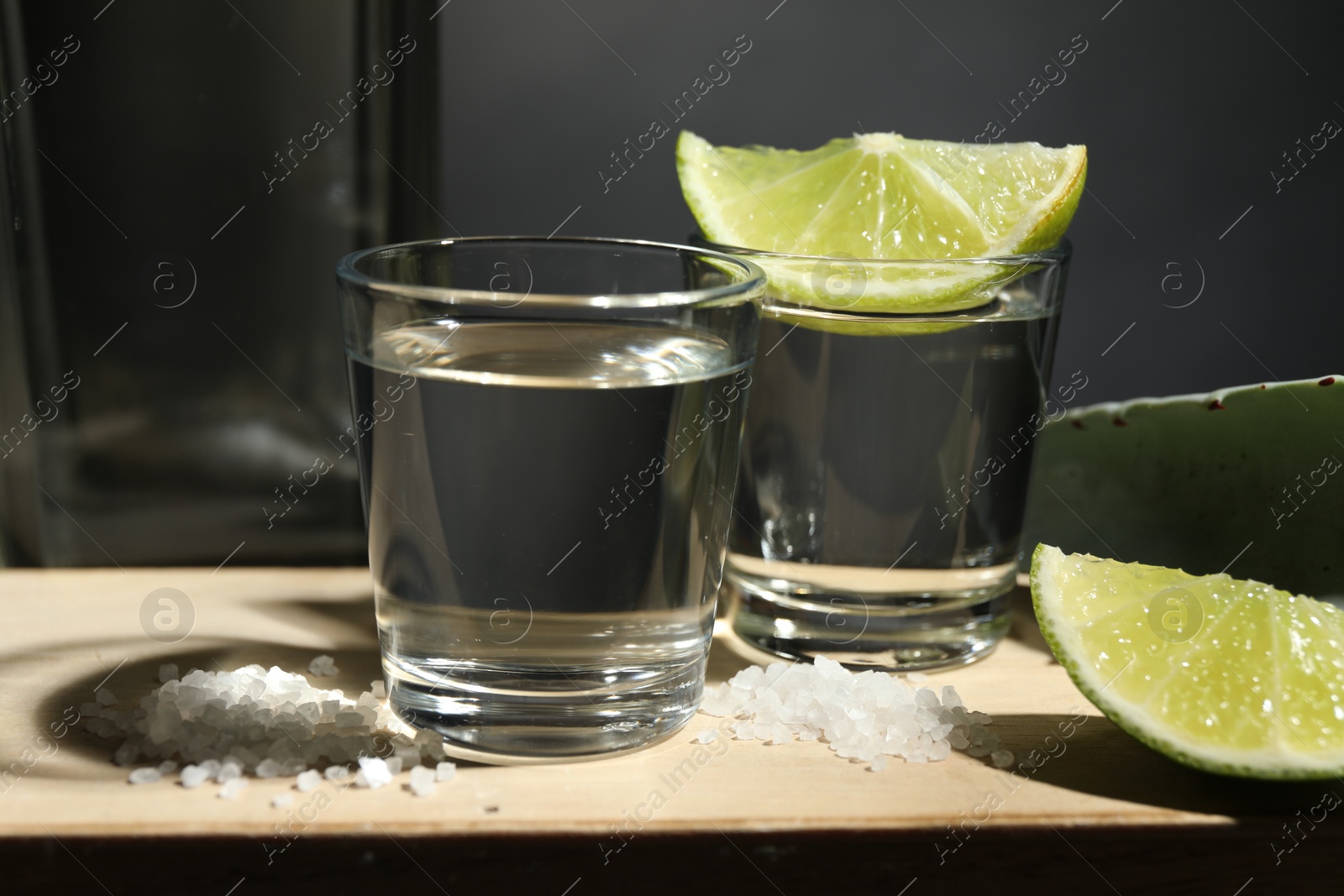 Photo of Tequila shots, slices of lime, salt and agave leaf on wooden table against black background, closeup