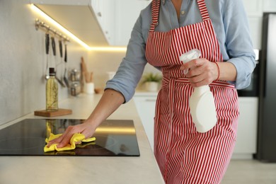 Photo of Woman cleaning induction cooktop with rag and detergent indoors, closeup