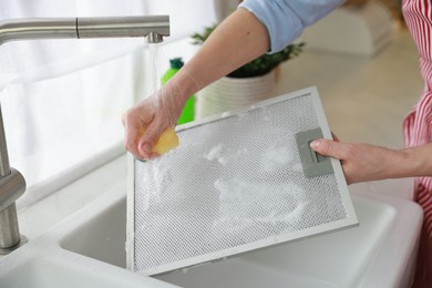 Photo of Woman cleaning filter of kitchen hood with sponge above sink indoors, closeup