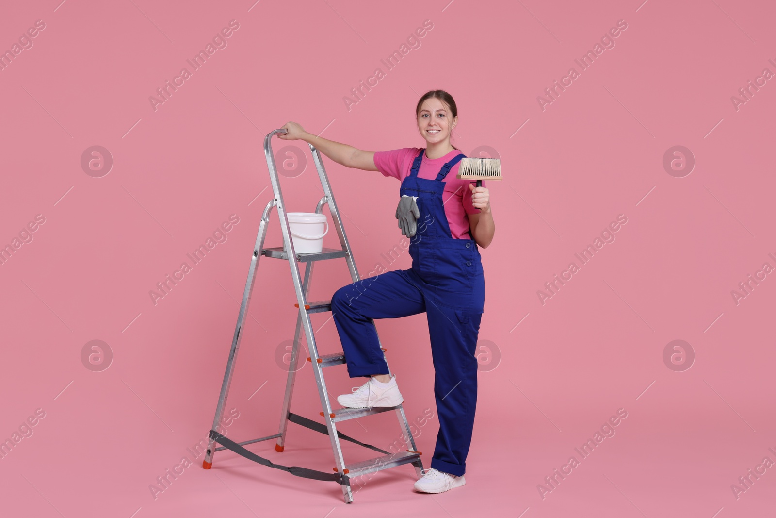 Photo of Professional painter with brush, bucket of paint and ladder on light pink background