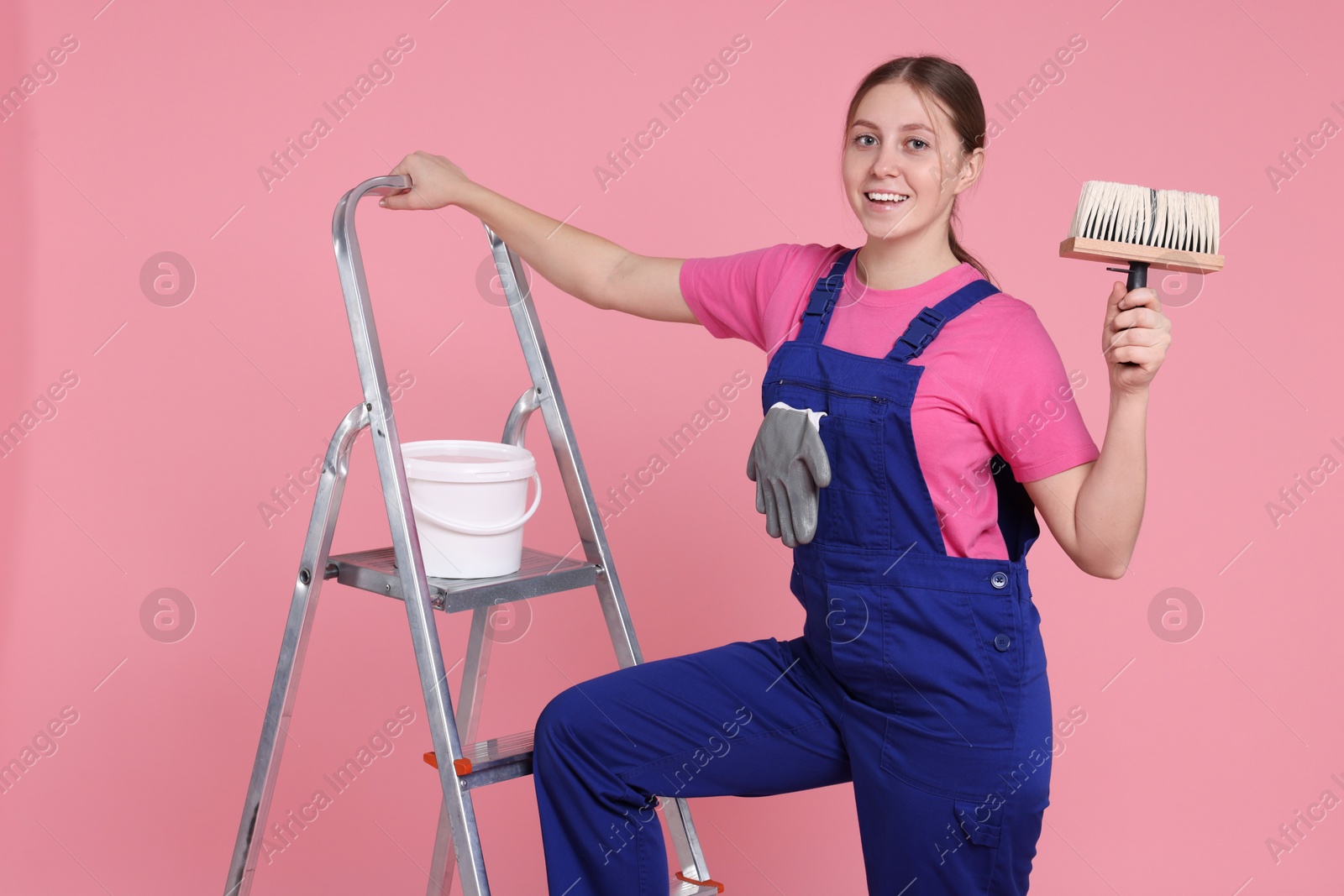 Photo of Professional painter with brush, bucket of paint and ladder on light pink background