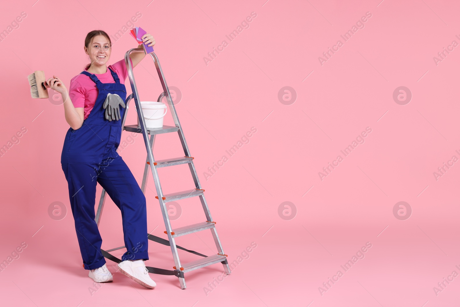 Photo of Professional painter with brush, color samples, bucket of paint and ladder on light pink background. Space for text