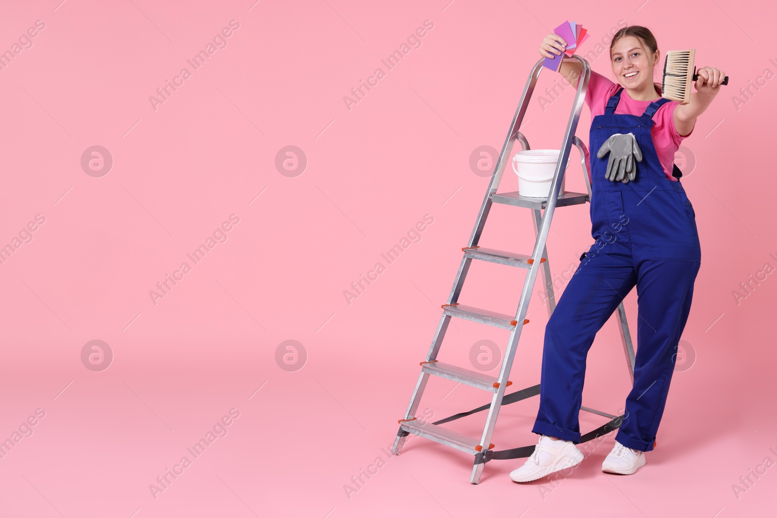 Photo of Professional painter with brush, color samples, bucket of paint and ladder on light pink background. Space for text