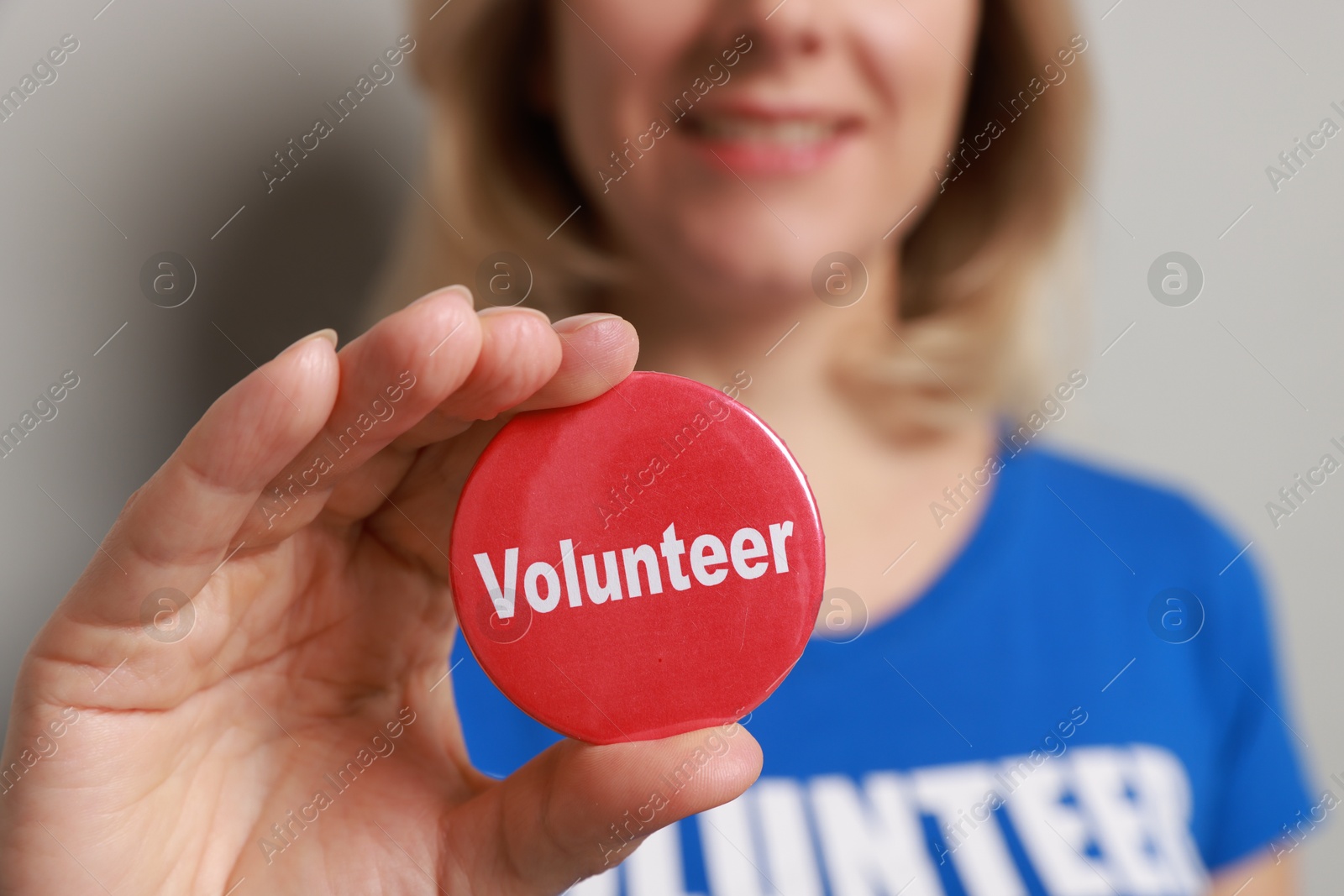 Photo of Woman holding button badge with word Volunteer on grey background, closeup