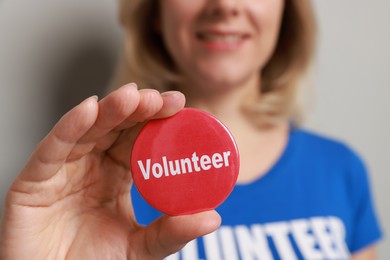 Photo of Woman holding button badge with word Volunteer on grey background, closeup