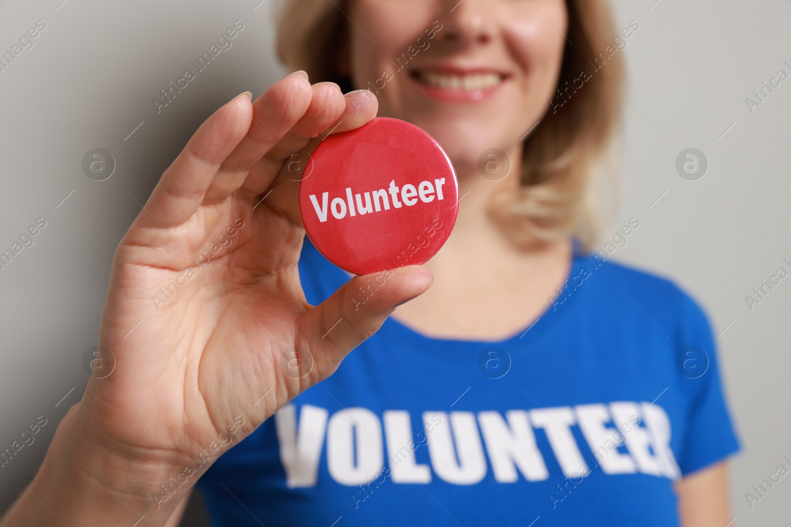 Photo of Woman holding button badge with word Volunteer on grey background, closeup