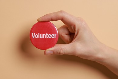 Photo of Woman holding button badge with word Volunteer on beige background, closeup