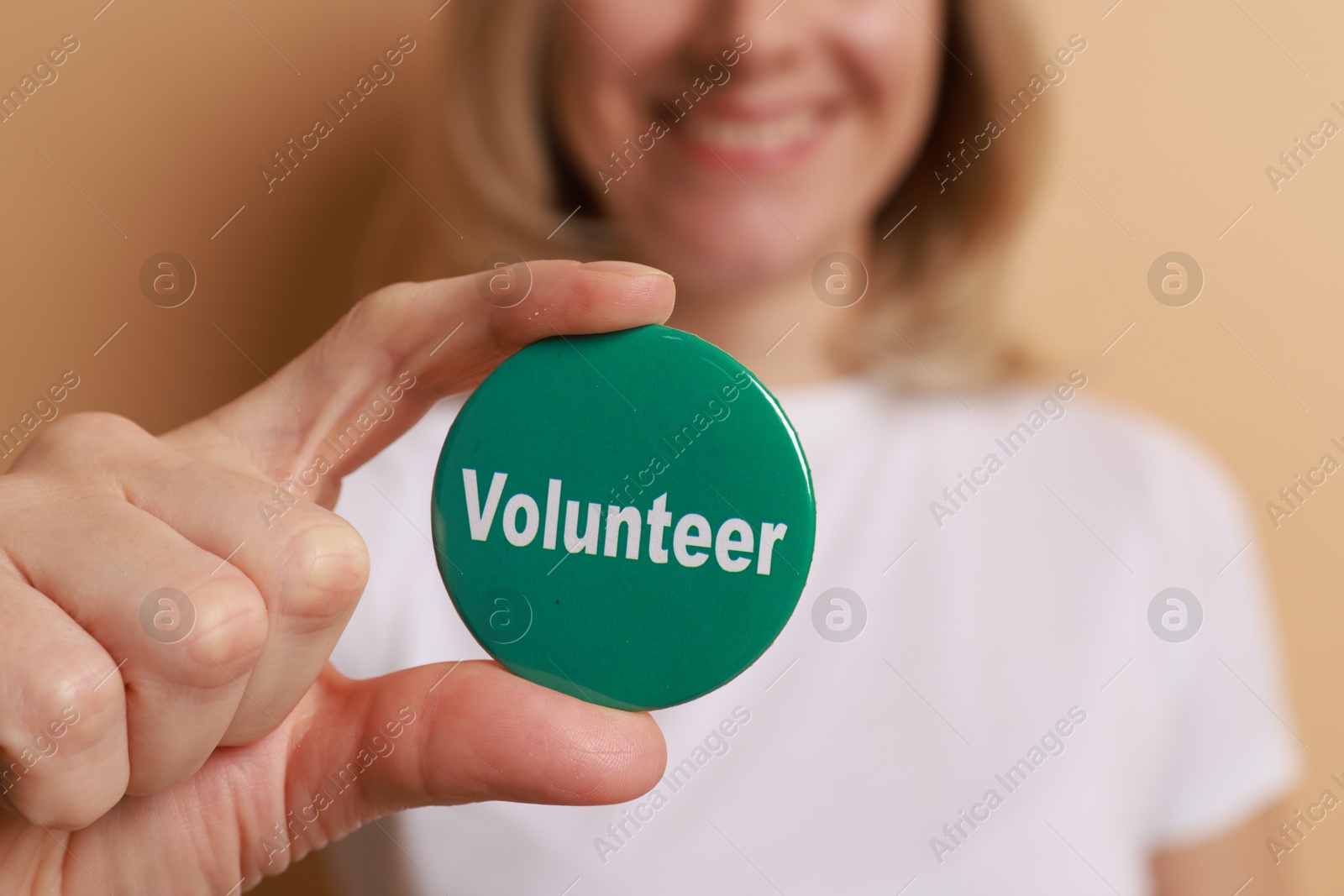 Photo of Woman holding button badge with word Volunteer on beige background, closeup