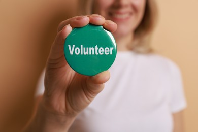 Photo of Woman holding button badge with word Volunteer on beige background, closeup