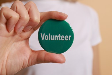 Photo of Woman holding button badge with word Volunteer on beige background, closeup