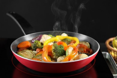 Photo of Frying pan with vegetables and stove on table against black background, closeup