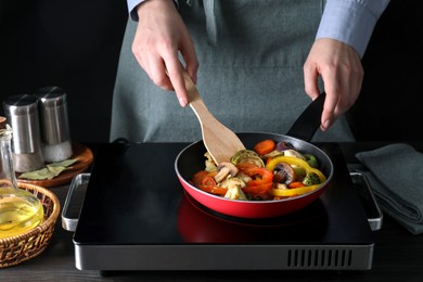 Photo of Woman frying vegetables and mushrooms in pan on stove at wooden table, closeup