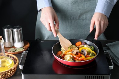 Photo of Woman frying vegetables and mushrooms in pan on stove at wooden table, closeup