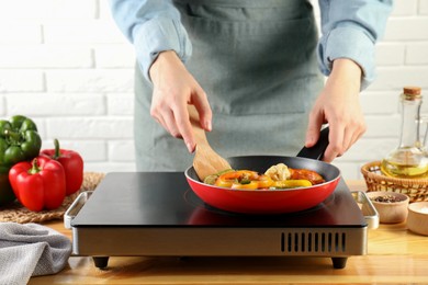 Photo of Woman frying vegetables in pan on stove at wooden table indoors, closeup