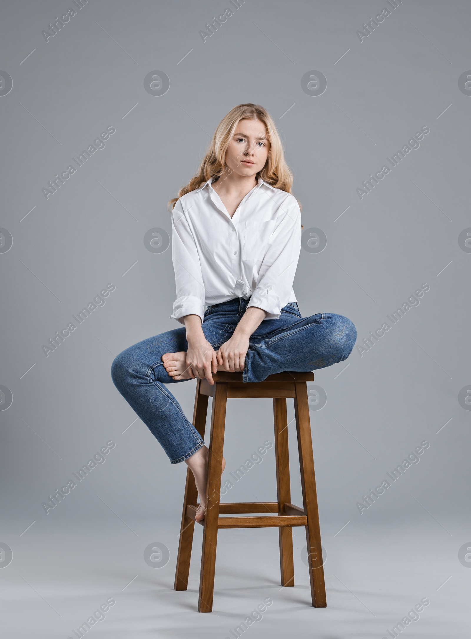 Photo of Beautiful young woman in stylish jeans sitting on stool against grey background