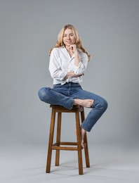 Photo of Smiling woman in stylish jeans sitting on stool against grey background