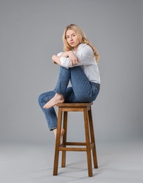 Photo of Beautiful young woman in stylish jeans sitting on stool against grey background