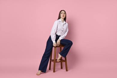 Beautiful young woman in stylish jeans sitting on stool against pink background