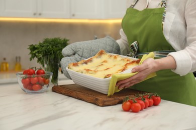 Photo of Woman putting baking dish with tasty lasagna onto white marble table in kitchen, closeup