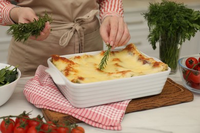 Photo of Woman putting rosemary onto tasty lasagna at white table indoors, closeup