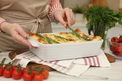 Photo of Woman putting rosemary onto lasagna at white marble table indoors, closeup