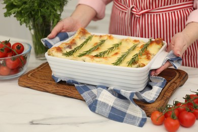 Photo of Woman putting baking dish with tasty lasagna onto white marble table indoors, closeup