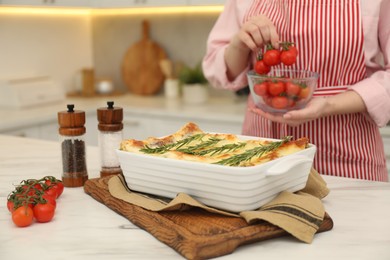 Photo of Delicious lasagna with rosemary on white marble table and woman with tomatoes in kitchen, closeup