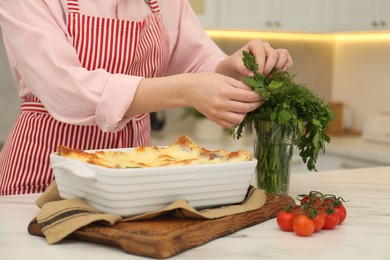 Photo of Woman taking parsley from glass near delicious lasagna at white marble table indoors, closeup