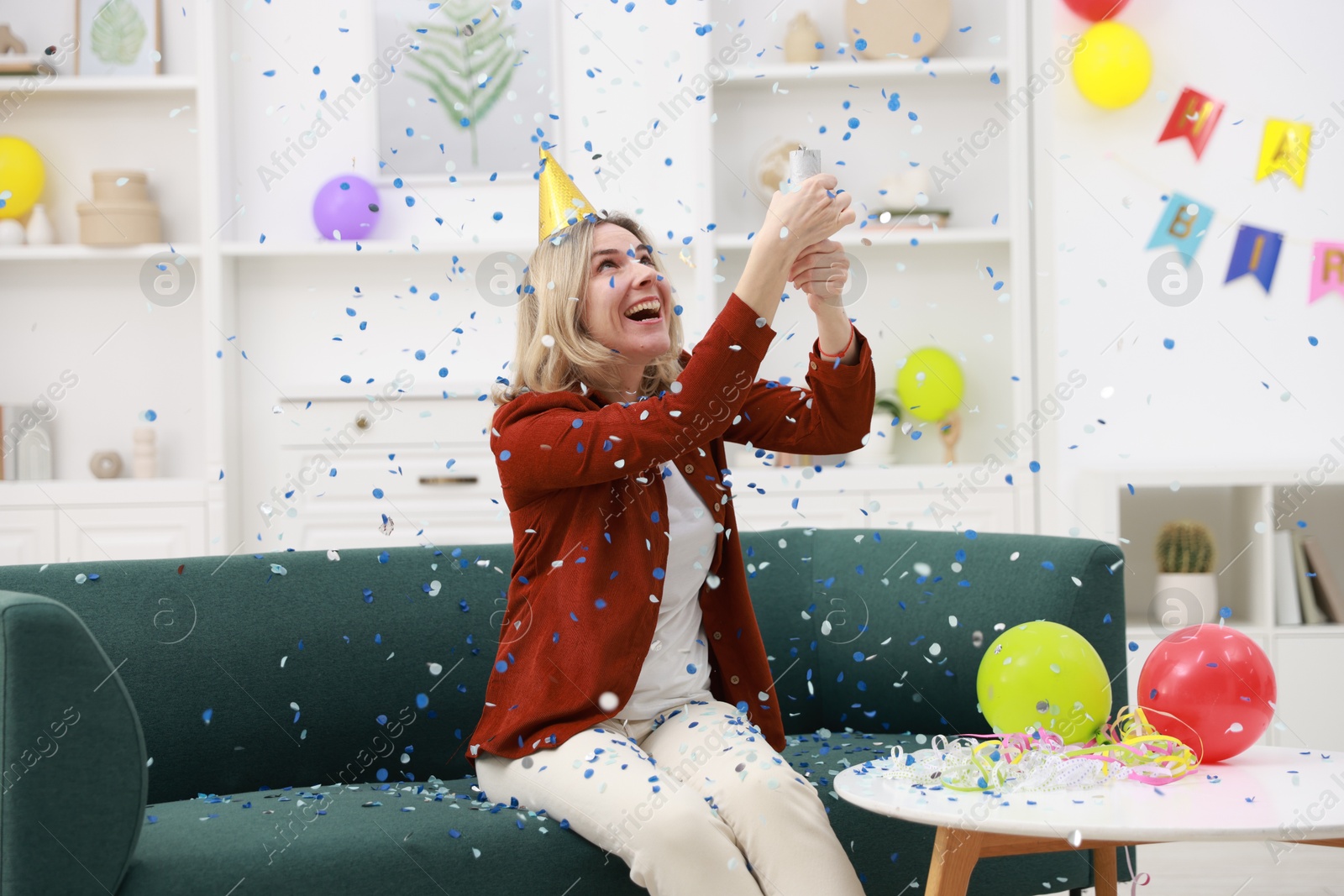 Photo of Happy woman blowing up confetti popper on sofa in room decorated for party