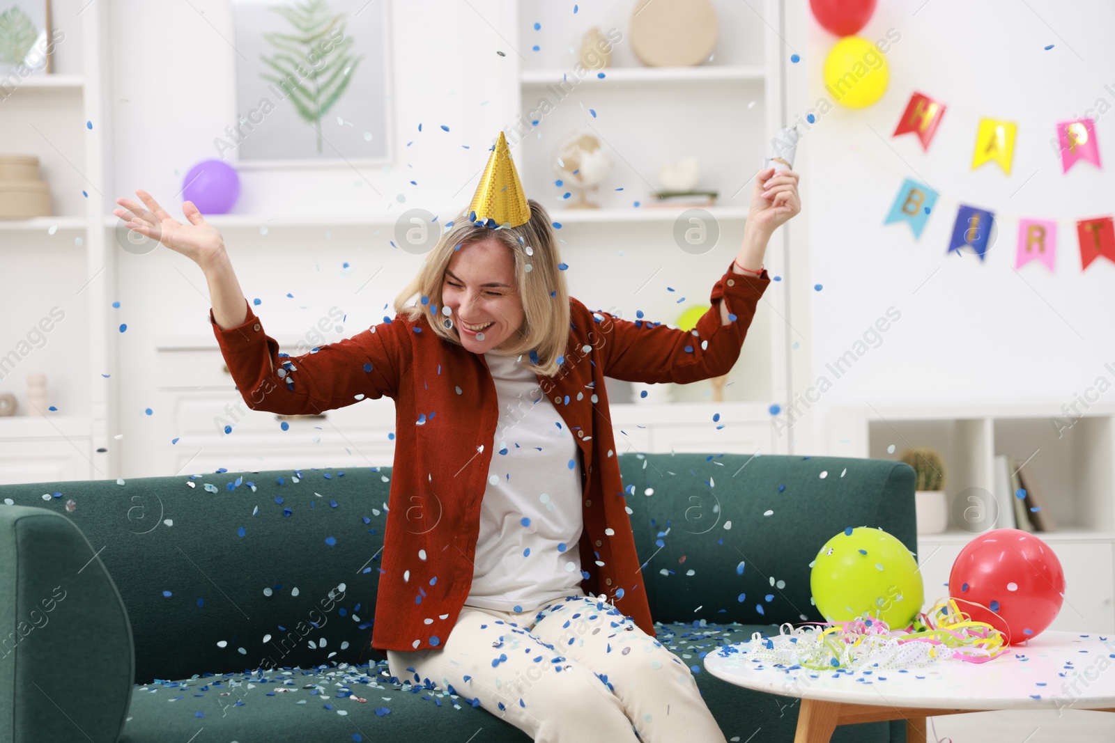 Photo of Happy woman blowing up confetti popper on sofa in room decorated for party