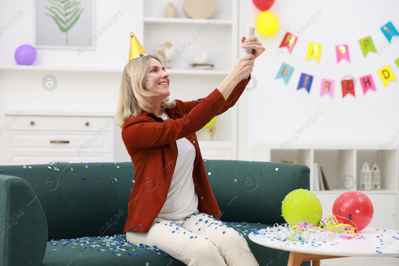 Photo of Happy woman blowing up confetti popper on sofa in room decorated for party