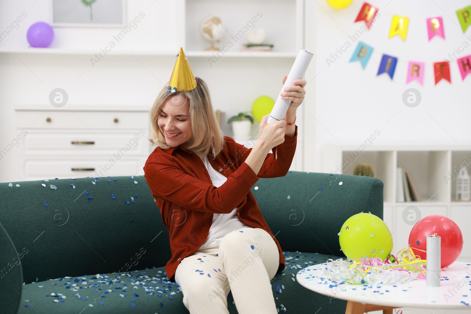 Photo of Happy woman blowing up confetti popper on sofa in room decorated for party