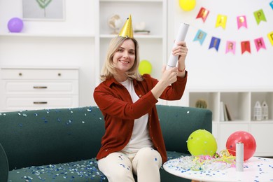 Photo of Happy woman blowing up confetti popper on sofa in room decorated for party