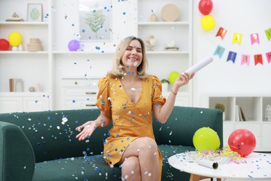 Photo of Happy woman blowing up confetti popper on sofa in room decorated for party