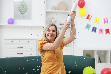 Photo of Happy woman blowing up confetti popper on sofa in room decorated for party