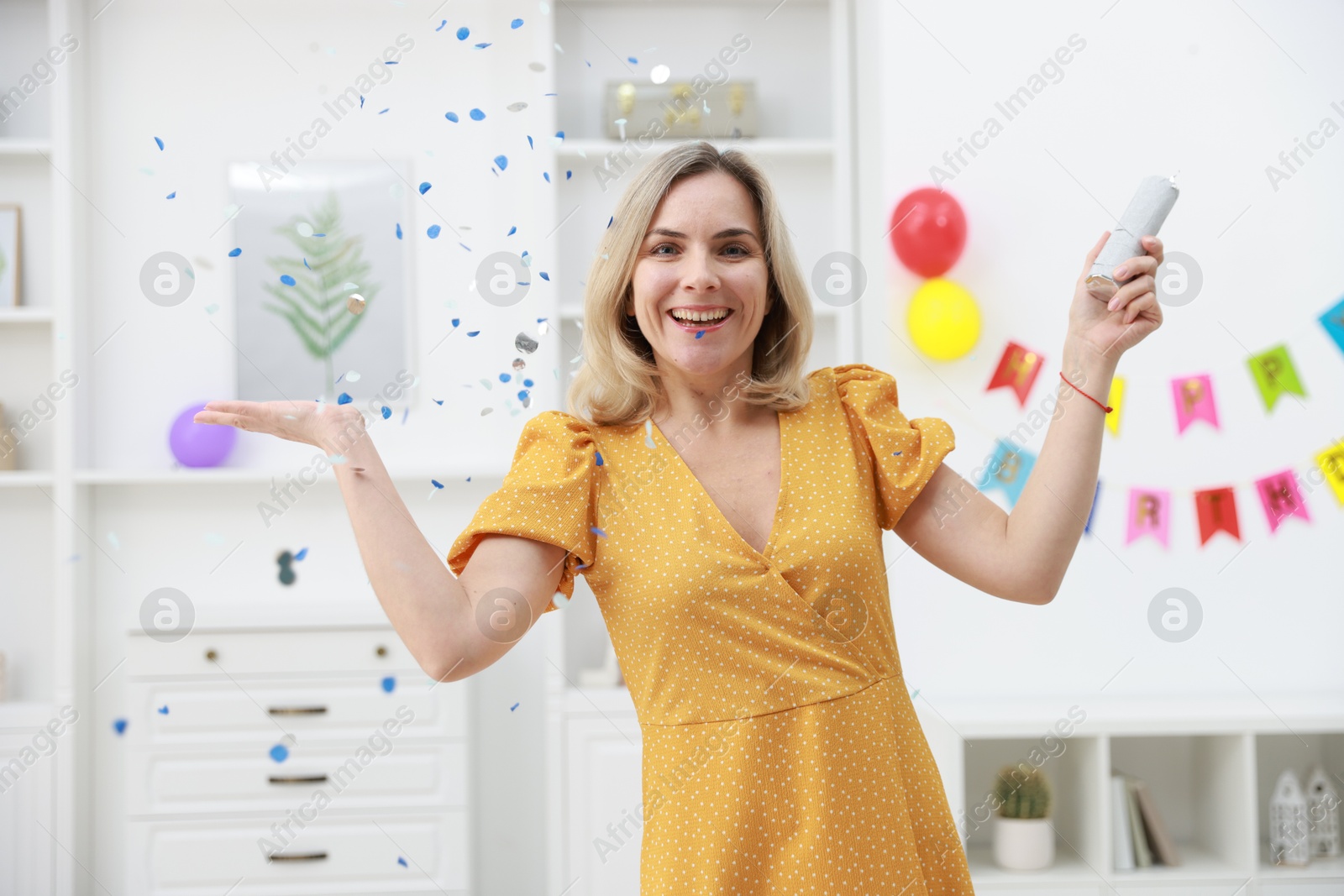 Photo of Happy woman blowing up confetti popper on sofa in room decorated for party