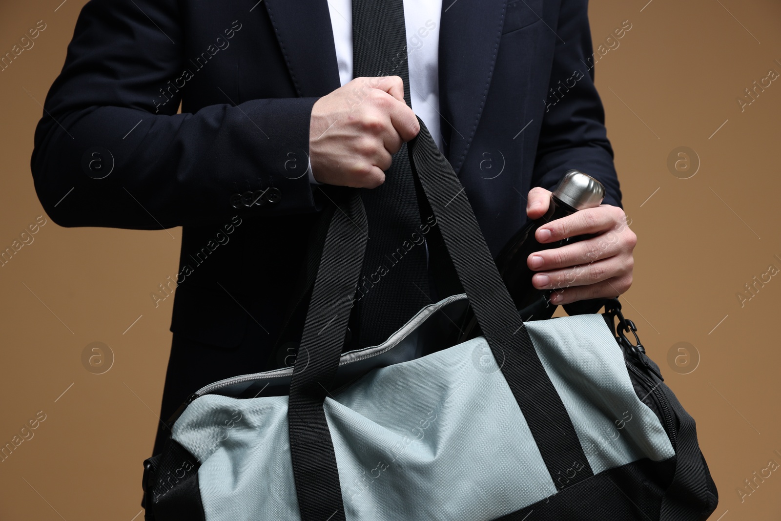 Photo of Man in suit putting bottle of water into gym bag on brown background, closeup