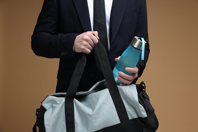 Man in suit putting bottle of water into gym bag on brown background, closeup