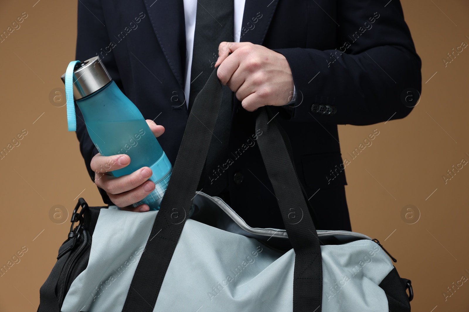 Photo of Man in suit putting bottle of water into gym bag on brown background, closeup