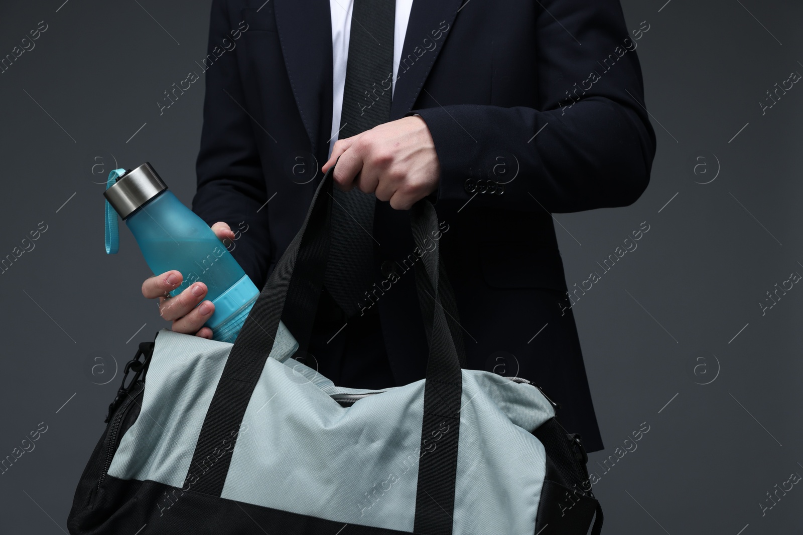 Photo of Man in suit putting bottle of water into gym bag on dark grey background, closeup