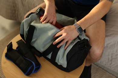 Photo of Man putting bottle of water into gym bag indoors, closeup