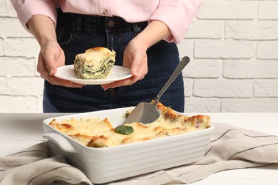 Photo of Woman holding plate with piece of delicious spinach lasagne at white wooden table, closeup