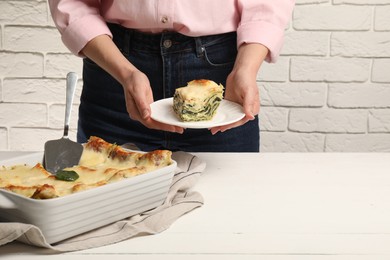 Photo of Woman holding plate with piece of delicious spinach lasagne at white wooden table, closeup